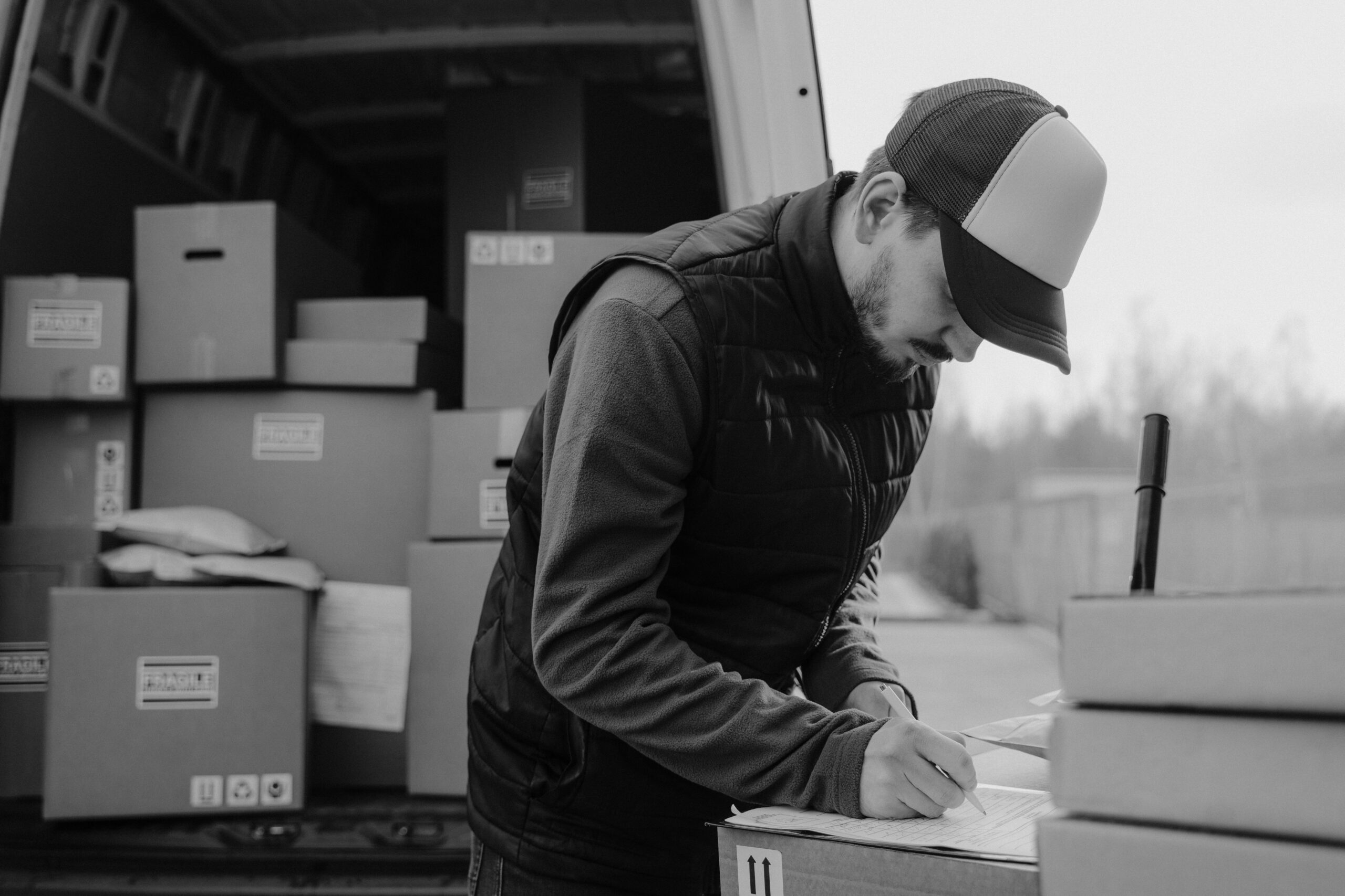 Black-and-white image of a courier documenting package details by a delivery van.